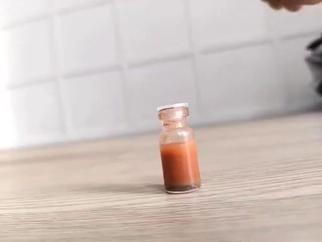 The video shows a close-up, low-angle shot of a tiny glass vial containing a reddish-brown liquid. The vial is standing upright on a light-colored, wooden surface. In the background, a white tiled wall is visible. A person's foot, specifically the toes and the side of the sole, is shown entering the frame from the right and moving towards the vial. The foot appears to be descending, suggesting an imminent action involving the vial. The lighting is bright, casting subtle shadows.