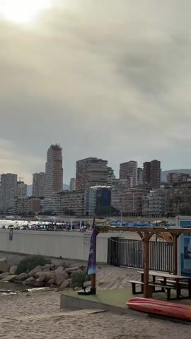 The video shows a beach with a city skyline in the background. The sky is cloudy with the sun peeking through. The beach is made of sand with some rocks near the water. There are several buildings in the background, including tall skyscrapers. There are some people on the beach and in the water. In the foreground, there is a picnic table with benches and a red kayak on the sand. The water is calm and the waves are small. There is a long concrete structure extending into the water. The overall scene depicts a coastal city on a cloudy day.