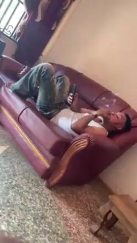 The video shows a man lying on a maroon leather sofa. He is wearing a white tank top and jeans. He is holding a smartphone in one hand and a lit cigarette in the other. He appears relaxed, looking at the phone. The sofa is placed in a room with tiled floors and a window visible in the background. A small wooden table is also present in the room.