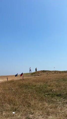 The video shows a wide shot of a grassy, hilly landscape under a clear blue sky. Two figures are visible on the crest of a hill, one appearing to be in a white shirt and the other in a dark shirt. In the middle ground, three golf bags are positioned on the left side of the hill. A white flag, likely a golf flag, is visible on the far right of the hill. The terrain is covered in dry, yellowish grass, with a few scattered green patches. A fence, consisting of thin posts and wire, runs across the middle ground of the image. The camera is static, capturing the scene from a low angle looking up towards the hill.
