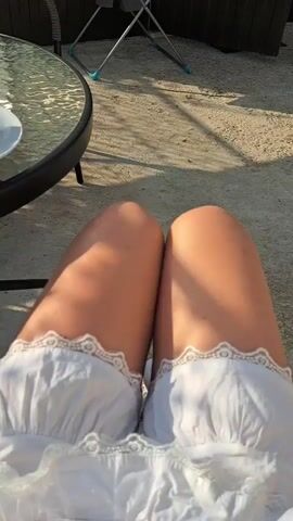 A person's legs are shown from the thighs down, wearing white lace-trimmed underwear and white knee-high socks. They are sitting outdoors, likely on a patio, with a glass-topped table to the left. In the background, there is a clothes drying rack with clothes on it and a blue basin. The sun is shining, casting shadows on the ground.