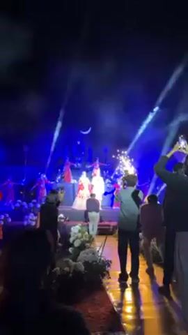 A wedding celebration unfolds under dramatic lighting, with the bride and groom standing on a stage as fireworks erupt behind them. The bride is adorned in a red and gold gown, while the groom wears a white outfit. Dancers in red dresses are also visible on the stage. The stage is set against a backdrop of a large blue Shiva statue, adding to the cultural significance of the event. The audience stands below, some capturing the moment with their phones. The ambiance is festive and celebratory.