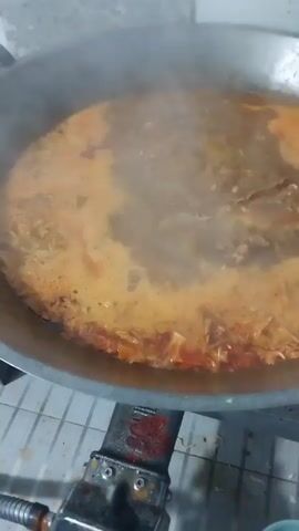 Here is a concise description of the video: 
The video shows a close-up shot of a large, silver wok-like pan on a gas stove. Inside, a bubbling orange-yellow liquid is simmering. The contents appear to be a seafood-based stew or soup, with pieces of what seems to be lobster being stirred with a metal spatula. Steam is rising from the hot liquid. The stove is an older model with a visible gas regulator. The background shows a white tiled wall and floor. The overall impression is of a cooking process in progress, likely in a commercial or large-scale cooking environment.