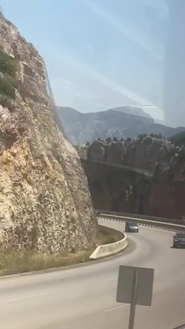 The video shows a winding road with cars and motorcycles driving on it. The road is bordered by a steep, rocky cliff face on the left and a guardrail on the right. In the background, there are mountains and a clear blue sky. The scene appears to be a sunny day in a mountainous region.