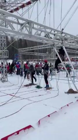 The video shows people working in a snowy field around a large white metal scaffolding structure, possibly for a training or obstacle course. Some individuals stand on the ground with ropes, while others are on the scaffolding, maneuvering with ropes and safety gear. The crowd of people nearby suggests an event or training session in progress. The setting is outdoors, with snow covering the ground and trees visible in the background.