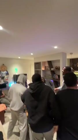 A group of about six men are in a living room, appearing to be celebrating or partying. They are gathered around a coffee table that has pizza boxes and beer bottles on it. One man in a gray sweatsuit is jumping up and down while the others are also excited. One man is sitting on a stool in the background. The room is well-lit and includes a built-in shelving unit and a TV.