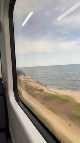 A view from a train window shows the ocean and the coastline. The train appears to be moving along the coast. The water is a deep blue, and the sky is partly cloudy. The coastline is rocky and has some vegetation.
