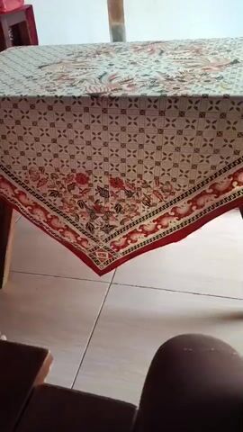 The video shows a close-up shot of a table covered with a batik tablecloth. The tablecloth has a geometric pattern and a floral border. The table is supported by wooden legs, and the floor is tiled. A dark object, possibly a piece of furniture, is visible in the foreground. The lighting suggests an indoor setting.
