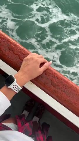 The media shows a person on a boat, holding onto the wooden railing. The person's hand with a white smartwatch is visible, along with their feet wearing leopard print slippers and a plaid shirt. The scene transitions to a wide shot of the ocean, with waves and foam indicating the boat is in motion. In the distance, a silhouetted island is visible against a sky with dramatic clouds and the sun peeking through, suggesting it is either sunrise or sunset. The water is a deep green, and the overall atmosphere appears serene and peaceful. The location is identified as Don Sak, Thailand. There is no visible text in the media.