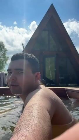 The video shows a man and a woman kissing in an outdoor hot tub in front of an A-frame building. The man is partially visible, revealing his upper back, shoulder, and the side of his head. The woman has reddish-blonde hair and is wearing a black swimsuit. The water in the hot tub is clear and appears to have jets creating some turbulence. The A-frame building behind them features large glass windows, providing a view of trees and other greenery inside. The sky is blue with scattered clouds. The sun appears to be shining brightly.