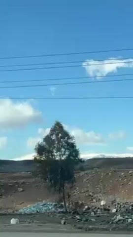 The video shows a landscape taken from a moving vehicle. The sky is blue with scattered white clouds. In the foreground, there is a metal utility pole with power lines stretching across the frame. The ground is rocky and barren, with patches of debris and sparse vegetation. In the distance, there are snow-capped mountains visible on the horizon. The camera pans upwards, emphasizing the sky and the power lines. The overall impression is a rural or industrial landscape on a sunny day.