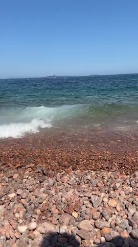 A POV shot shows a pebble beach with a person swimming in the ocean. The water is turquoise and clear. The sky is a bright blue, with a distant landmass. The camera pans slightly to the right, showing a few more waves.