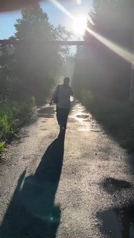 A person is walking away from the camera on a paved path. The sun is shining brightly from behind the person, creating a strong silhouette and lens flare. There are puddles on the path, indicating recent rain or moisture. Trees and foliage are visible on either side of the path, and a large pipe or structure runs overhead in the background. The person is carrying a bag over their shoulder. The overall atmosphere is serene and bathed in golden light.