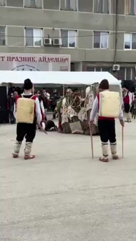 The video shows a group of traditionally dressed people performing a ritual dance in a town square. Two figures, seen from behind, wear black pants, white shirts, red vests, and a large square, straw-like object strapped to their backs. They also wear traditional Bulgarian shoes and hats. They hold sticks that they periodically strike together, creating a rhythmic sound. A large figure draped in layers of fabric and adorned with various objects moves in the center of the group. Other people, also in traditional clothing, join in the dance, including women in colorful skirts and vests. In the background, a building with multiple windows stands behind a tent. There are balloons in the colors of the Bulgarian flag: white, green, and red. The overall scene suggests a celebration or festival rooted in Bulgarian tradition. A banner is visible above the tent, but the text is illegible.
