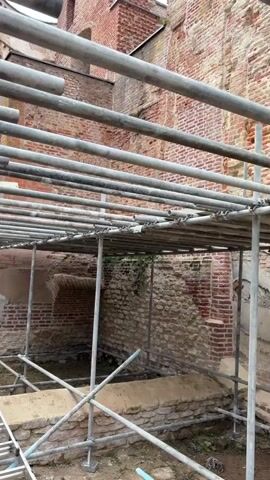 The video shows a construction site with scaffolding and a ladder. The scaffolding is made of metal pipes and supports. A ladder is leaning against a wall. The walls are made of brick and stone. There is some vegetation growing on the walls. The ground is covered in dirt and debris. The scene appears to be outdoors. There are no people visible in the video.