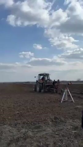 The video shows a maroon and green tractor plowing a field. The tractor is in the middle of the frame, moving from right to left across a field of dark brown soil.  Behind the tractor, the plowed soil is visible, with a tripod set up on the edge of the field. The sky is blue with scattered white clouds. In the distance, a slightly hazy skyline can be seen.