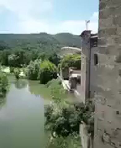 The video shows a wide, slow-moving river with green-tinted water, flowing through a valley surrounded by lush green trees. The trees extend up the slopes of rolling hills in the background. On the right side of the frame, a section of a stone building is visible, appearing to be an old structure, possibly a castle or an old wall, with some vegetation growing around it. The sky is blue with a few scattered clouds. The camera appears to be mounted on a moving object, possibly a drone or a vehicle, as the perspective shifts slightly throughout the short clip, giving a dynamic view of the landscape. There are no people or animals clearly visible in the video. The overall impression is a peaceful, natural scene with a hint of historical architecture.
