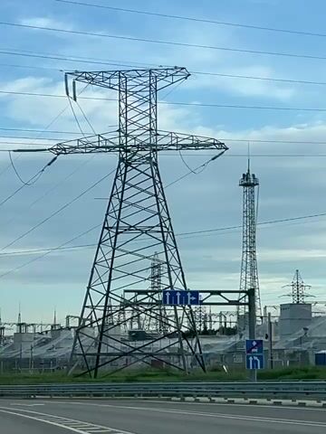 The video shows an electrical substation filmed from a moving vehicle. The vehicle moves along a road marked with lane direction signs, passing by a large metal electricity pylon. Behind the pylon and the signs, the substation is visible, featuring numerous transformers, supporting structures, and additional pylons. The sky is blue with patches of white clouds.