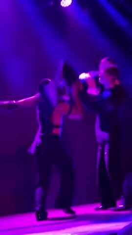 Here's a description of the video:

The video shows a performance on a stage lit with vibrant blue and purple lights. Three performers are visible. A dancer is wearing black lingerie-style clothing, including what appears to be a bra, and long gloves. This performer interacts with two other dancers wearing similar, mostly-black, costumes. One of the dancers is briefly lifted. The environment is dark and hazy, typical of a concert or club atmosphere.