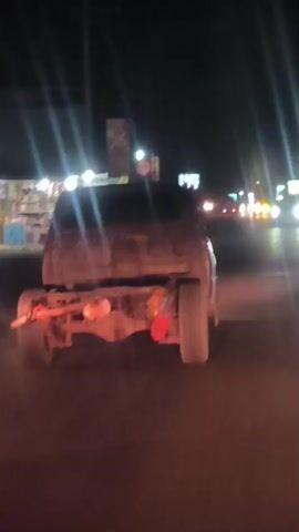 The video captures the rear view of a vehicle's chassis at night. The main subject is what appears to be the stripped-down frame of a truck or utility vehicle, lacking its main body or bed. Visible components include the rear wheels, axle, and suspension. Two prominent orange/red spherical objects, possibly gas cylinders or tanks, are attached to the chassis, along with a red bag or bucket near the right rear wheel.

The setting is outdoors at night, indicated by the darkness and numerous artificial lights. In the background, brightly lit commercial establishments or roadside stalls are visible on the left, and distant streetlights or traffic lights can be seen along a road. The scene suggests an urban or semi-urban environment in Lancokpanteara, Indonesia.

A sign with the number "76" and the word "APEL" (possibly a brand or product, with "INDONESIA" partially visible below) is discernible in the background. Another sign shows the number "50", which could indicate a speed limit. The vehicle appears to be stationary or moving slowly, and the video is likely filmed from another vehicle.
