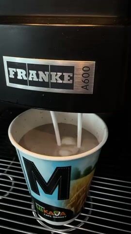 The video shows a close-up of a drink being dispensed into a paper cup. The cup is branded with the logo "M LITAKA CAFE MARKET" and features a scenic image. The drink appears to be a coffee-based beverage, possibly a latte or cappuccino, with milk being frothed and poured into the cup.

Above the cup, a black appliance with the "FRANKE A600" logo is visible, suggesting it's a commercial coffee machine. The setting is indoors, likely a cafe or market, given the branding on the cup. The lighting is artificial, and there are no indications of the time of day or weather. The focus is entirely on the beverage preparation process.

The video captures the dynamic motion of the milk swirling and mixing with the coffee, creating a foamy texture. The stainless steel grate beneath the cup adds to the impression of a professional setting. The overall mood is one of everyday convenience and the simple pleasure of a freshly made hot drink.