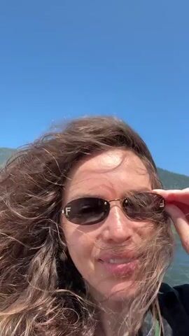 A person wearing sunglasses is on a boat. Their long, brown hair is blowing in the wind. The person is smiling and adjusting their sunglasses with their right hand. Behind them, a body of water is visible, with a forested mountainside in the distance under a clear blue sky.