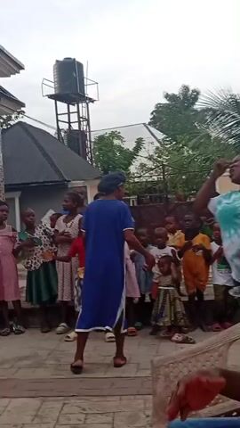 The video depicts an outdoor gathering, likely a children's party or celebration, in what appears to be a residential courtyard or backyard in Onicha, Nigeria. Numerous people are present, including many children and several adults.

In the initial frames, a woman holding a child in a red dress is seen singing or shouting enthusiastically, surrounded by other children who are looking on. A large water tank on a stand is visible in the background, along with lush green foliage and palm trees. The scene then pans to show a modern-style house with a grey roof and brick detailing, and more children and adults gathered. A table laden with drinks and snacks is set up, suggesting a party atmosphere.

Further into the video, children are seen standing and watching, while adults are seated on plastic chairs. One young boy in an orange shirt is standing in the foreground, seemingly addressing the group or performing. The overall mood appears joyful and relaxed. The time of day seems to be daytime, possibly late afternoon, with soft, diffused lighting. A child in the background wears a white t-shirt with "GUCCI" written on it.