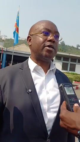 A man in a suit and glasses is being interviewed outside. He is speaking into a Sony recording device held by a person whose hand and part of their arm is visible. The man is bald and wearing a white shirt under a dark jacket. Behind him, a building and the flag of the Democratic Republic of the Congo are visible. The sky is overcast.