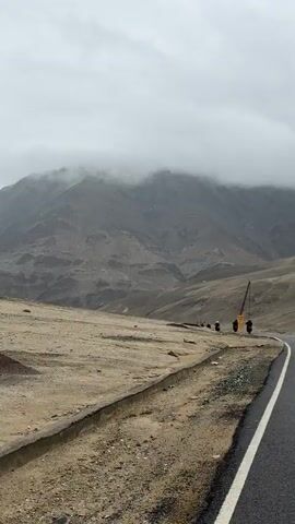 The video shows a winding asphalt road with a white line down the center, curving towards the right of the frame. The road passes through a barren, dusty landscape with sparse vegetation. In the distance, there are several figures on motorcycles, appearing to be traveling along the road. A crane is visible near the road. Behind the landscape, a large, mist-shrouded mountain range dominates the background. The sky is overcast and cloudy, suggesting a cool or damp climate.