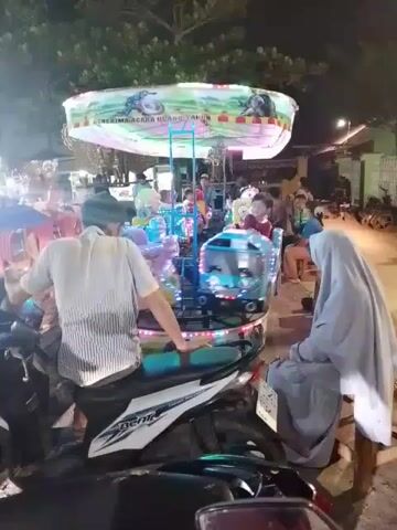 The video shows a small merry-go-round operating in an outdoor setting at night. The merry-go-round has various colorful seats shaped like cartoon characters. Several children are riding on it. A man wearing a white shirt and cap is sitting on a scooter parked directly in front of the merry-go-round, facing away from the camera, with his arm resting on the scooter’s seat. To his right, a woman wearing a grey hijab is sitting on a chair. Several other people are standing or sitting around the merry-go-round. There are some trees, buildings, and streetlights visible in the background. The sign at the top of the merry-go-round reads "Menerima Acara Ulang Tahun."