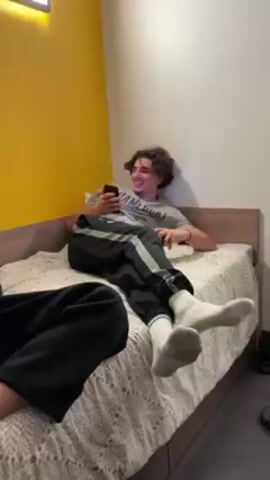 Two young men are on a bed in a room with a yellow accent wall and a white wall. One man is propped up against the headboard, looking at a phone in his left hand and holding a blue video game controller in his right hand, smiling. He is wearing a grey t-shirt, black track pants with grey stripes, and white socks. The second man is sitting cross-legged at the foot of the bed, looking at his own phone. He is wearing a green long-sleeved shirt and black sweatpants. The bed has a light-colored patterned bedspread.