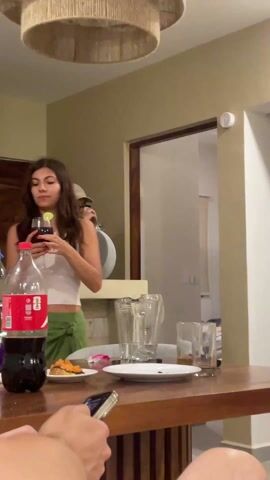 The video shows a woman standing by a wooden table with a large bottle of Coca-Cola and a wine glass. She takes a sip from the glass, which contains a dark red liquid and a lime wedge. Another person stands behind her, and a third person is partially visible at the edge of the frame, holding a phone. The table has plates with leftover food, a blender, and empty glasses on it. There is a doorway behind the table, and a light fixture hangs from the ceiling. A speaker sits on the counter behind the table. The video appears to have been taken indoors in a casual setting with background music playing.