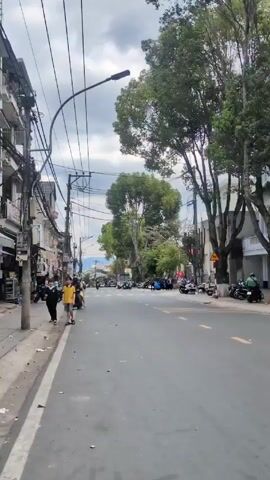 The video shows a street in a town with a cloudy sky overhead. On the left side of the street, there are buildings with exposed wires overhead, a sidewalk, and litter. Two young people are walking on the sidewalk. The street has a few vehicles on it, including motorcycles. On the right side of the street, there are trees, buildings, and more motorcycles. People can be seen standing or sitting in the distance.