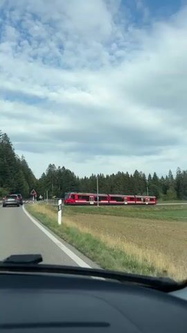 A red and white passenger train travels on tracks to the right of a paved road. The road is lined with white and black striped posts. A dark colored SUV is driving on the road in the same direction as the train. The road is bordered by a grassy field. In the background, a dense forest of green trees stretches across the horizon. The sky above is blue with scattered white clouds. A sign with a red triangle is visible by the side of the road. The video is captured from inside a vehicle, likely from the dashboard or windshield.