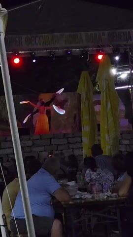 This media file depicts an outdoor evening event, likely a festival, at Gara Oreshets, Bulgaria. The scene is set at night, illuminated by stage lights and ambient lighting. A performer is visible on a stage, dressed in a yellow skirt and a dark top, manipulating what appear to be glowing, fan-like props. The stage is backed by a screen displaying an image and above it, a banner with Cyrillic text is visible. The text appears to read "ОРЕН ФЕСТИВАЛ "ЖИВА ВОДА" ВЕНЕЦИЯ", which translates to "OREN FESTIVAL "LIVING WATER" VENICE".

In the foreground, several people are seated at tables, suggesting a dining or gathering area associated with the festival. They are casually dressed and appear to be enjoying the performance and the atmosphere. Yellow umbrellas are set up, likely for shade or shelter, and large trees with dark foliage frame the upper part of the scene. The ground is covered with gravel and some patches of grass. The overall mood seems relaxed and festive, with the glowing lights of the performance adding a touch of magic to the night. A white tent structure is also present on the left side of the frame, further indicating an organized event. Flags are visible on a pole in the background, hinting at a national or cultural gathering.