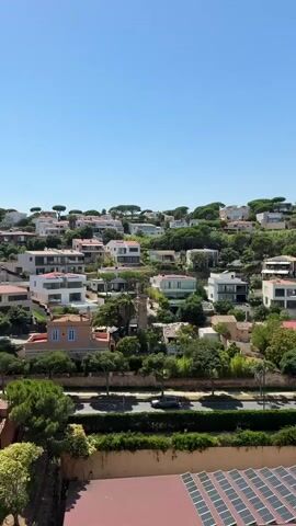 This is a vertical video taken from a high vantage point, looking down on a residential area on a hillside. The scene is bathed in bright sunlight under a clear blue sky, indicating it's daytime, likely late morning or early afternoon. Lush green trees and manicured hedges are interspersed among the houses, many of which are modern with white facades and flat roofs, while others have traditional terracotta tile roofs. Some houses feature swimming pools and solar panels. A road winds through the neighborhood, with a few cars visible. In the foreground, there's a red-tiled roof with solar panels. The overall impression is one of a well-maintained, affluent community in a Mediterranean climate. There is no visible text in the video.