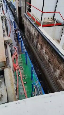 The video shows a towboat navigating through a lock. The boat is long and narrow, with a green deck and blue railings. A thick red rope is coiled on the deck. The towboat is moving through a narrow channel between two concrete walls. Water is swirling around the boat, indicating movement. The lock walls are textured and appear to be made of stone or concrete. There are white structures with red railings visible at the top of the lock walls. The sky is overcast. The video provides a close-up, high-angle view of the boat and the lock.
