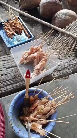 The video shows a close-up of a person preparing skewers of raw chicken. The person is holding a skewer of chicken in their left hand and a brush in their right hand, which they are using to coat the chicken with a dark sauce. In the background, there are more skewers of raw chicken, a blue tray, a plastic container, and a red woven tray. There are also coconuts and a broom visible. The setting appears to be outdoors, with a dirt ground and a wooden structure in the background. The video focuses on the preparation of the food, with no visible interactions between people.