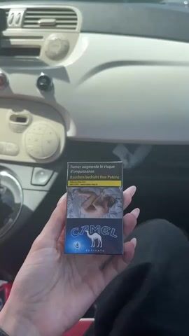 The video shows a close-up of a person's hand holding a pack of Camel Activate cigarettes. The hand has long, manicured fingernails with white French tips. The cigarette pack is dark blue with an image of a camel and text in French and German. The background appears to be the interior of a car, with the dashboard and steering wheel visible. The lighting suggests it is daytime. The video is short, with the camera slowly rotating the cigarette pack.