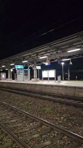 The video shows a view of a train platform at night. The platform is covered by a metal roof with lights illuminating the area. Train tracks are visible along the side of the platform. Signage indicating platform numbers is present. A person walks by in the latter part of the video.