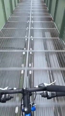 A first-person perspective video shows a person cycling across a metal bridge. The bridge's floor is constructed from longitudinal metal grates, and the rider's bicycle handlebars and front wheel are visible in the foreground. The camera then pans right, revealing the bridge's green metal girder structure. Beyond the bridge, a wide river stretches out, with lush green trees lining the opposite bank. The sky above is overcast, and several yellow buoys are visible in the river. The scene depicts a tranquil daytime ride over a river, likely the Main River in Kelsterbach, Germany.
