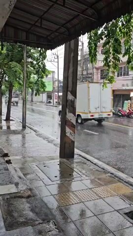 The video shows a wet street with a sidewalk in the foreground. Rain is falling, and the road and sidewalk are glistening. Several buildings line the street, with awnings and signs visible. A few motorbikes and a pickup truck are parked or moving on the street. Trees with green leaves are visible, some with branches hanging over the sidewalk. The scene appears to be an urban or town setting during a rain shower.