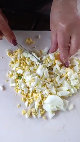 A close-up shot shows a person's hands chopping hard-boiled eggs on a white cutting board. The eggs are already chopped into various sizes, with distinct yellow yolks and white egg whites. One hand holds a knife, while the other hand is seen near the eggs, possibly steadying them. The background is dark and out of focus. The lighting suggests an indoor setting. The scene is likely part of a food preparation process, possibly for a salad or another dish. There is no visible text in the media.