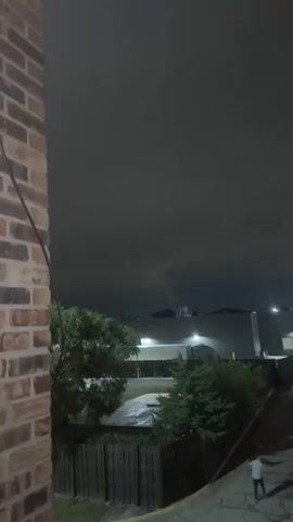 A brick wall is visible on the left side of the frame. Beyond the wall, there is a nighttime scene with a dark, cloudy sky. In the distance, several buildings are illuminated by artificial lights. There are trees and a wooden fence in the mid-ground. A paved area with a slight slope is visible in the foreground on the right, and a person is walking on it.