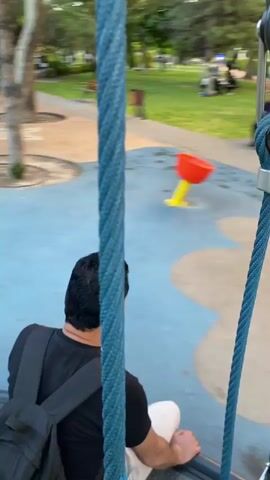 A person with a backpack is sitting on a swing in a playground. The playground has blue and pink surfacing with yellow poles. In the background, there are trees, buildings, and other people. The camera is moving, creating a blurred effect.