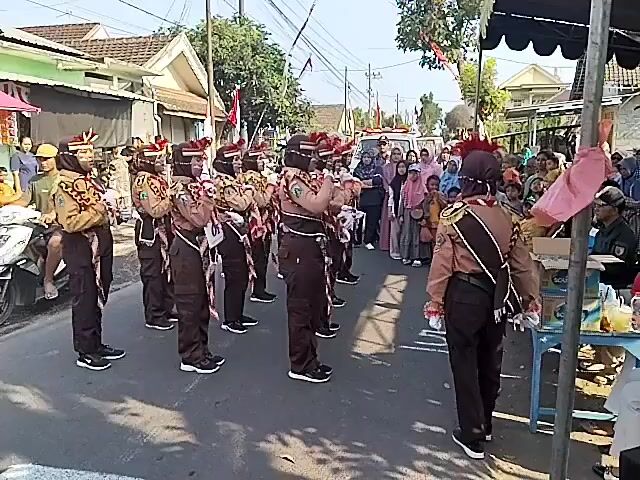 The video shows a group of scouts performing in a street parade. The scouts are dressed in brown uniforms with red and white sashes and feathered hats. They are holding batons and appear to be marching or performing a drill. There are spectators on the side of the street, some of whom are holding flags. The background shows houses and trees, indicating an outdoor setting. The weather appears to be sunny. The video is shot from a slightly elevated angle. The sound in the video includes music and cheering.