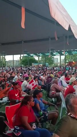A wide shot captures a large outdoor gathering under a canopy structure. A crowd of people, diverse in age and gender, are seated on the ground and in chairs, facing an unseen focal point. Many individuals are wearing casual clothing, with some displaying red and white attire, potentially indicating support for a team or event. The atmosphere appears relaxed and engaged. In the background, a building with multiple windows and a fence suggest an urban or suburban setting. The lighting indicates it is either late afternoon or early evening. The camera pans slowly, revealing the extent of the gathering and the various individuals within it.