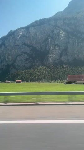 This video captures a side-view of a scenic landscape from a moving vehicle. The foreground shows a blur of a road with white and gray lines, indicating speed. Beyond the road, there's a bright green grassy field bordered by a light-colored metal guardrail. In the middle ground, a line of trees with full green canopies runs horizontally across the frame. Further back, the landscape transitions to a dense green forest that climbs the lower slopes of a massive, rugged mountain. The mountain itself is characterized by its sheer, rocky gray face, with visible striations and shadows suggesting its immense scale and geological formation. The sky above is a clear, bright blue, indicating a sunny day. The movement of the vehicle is evident in the motion blur of the foreground and the slight shift in the perspective of the trees and mountain. A dark-colored car is visible passing by in the middle distance, further emphasizing the sense of travel. The overall impression is one of a swift journey through a majestic natural environment.