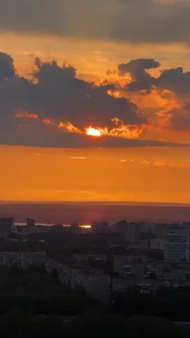 The video captures a picturesque sunset over a city, likely taken from a high vantage point. The sky is dominated by vibrant orange hues, with dark, dramatic clouds partially obscuring the setting sun. The sun itself appears as a bright, glowing orb, peeking through breaks in the clouds. Below the horizon, a wide expanse of orange light stretches across the sky, with a visible reflection of the sun on a distant body of water or reflective surface.

In the foreground and midground, a sprawling cityscape is visible, with numerous buildings and residential blocks silhouetted against the bright sky. Patches of dark green trees are interspersed among the buildings, suggesting urban greenery. The overall mood is serene and awe-inspiring, highlighting the natural beauty of the sunset contrasting with the urban environment. The clouds slowly shift throughout the video, changing the appearance of the sun's visibility. There are no people visible, and no discernible text. The time of day is clearly sunset or late evening, as indicated by the position and color of the sun. The setting appears to be an urban area in Bashkultaevo, Russia, based on the provided context.