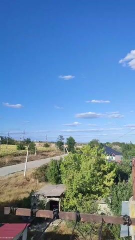 This is a vertical video of an outdoor scene on a clear day with a bright blue sky dotted with a few white clouds. In the foreground, there is a rusted metal railing with some black tape wrapped around it. Below the railing, partially visible, is a red roof on a building. Further back, there's an old, dilapidated structure with a dark opening. A road curves through a landscape of dry grass, green trees, and fields. In the background, houses with dark roofs are visible amongst the trees. Power lines and poles run along the side of the road. The overall impression is a rural or semi-rural environment.